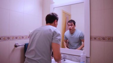 Adam banyo aynasında kendine bakarak yüzünü yıkıyor ve kurutuyor.