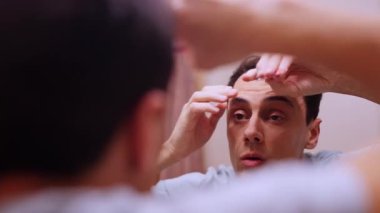 Adam kaşlarını ve burun kıllarını banyodaki aynanın önünde cımbız ve elektrikli budayıcı kullanarak koparıp buduyor.