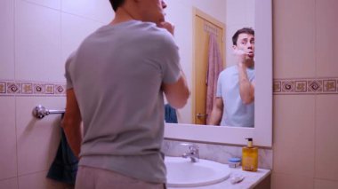 Adam dişlerini fırçalıyor ve banyo aynasında yüzünü yıkıyor.