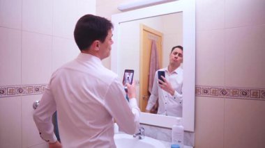 Narsist işadamı banyo aynasında akıllı telefonuyla fotoğraf çekiyor. Saçını ve kıyafetlerini düzeltiyor.