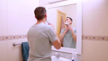 Beyaz bir adam banyodaki aynada sakalını kesiyor.