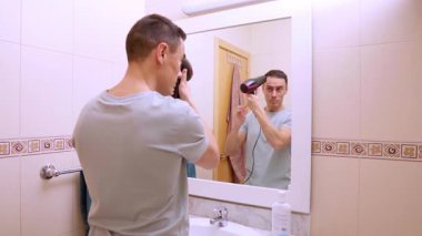 Adam banyodaki aynaya bakarken saç kurutma makinesi kullanıyor, özel bakım ve bakım rutinine odaklanıyor.