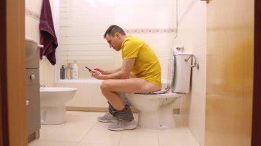 Yetişkin bir adam tuvalette otururken akıllı telefonunu kullanıyor.