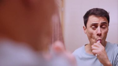 Yetişkin bir adam dişlerini fırçalar ve banyo aynasında kendine bakar.