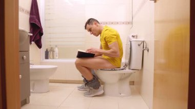 Modern bir banyoda tuvalette oturmuş kitap okuyan konsantre bir adam.