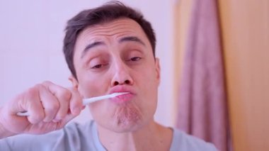 Yetişkin bir erkek dişlerini fırçalıyor. Banyodaki aynadaki yansımasına bakıyor.