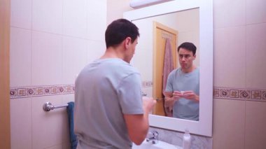 Adam banyo aynasının önünde ağız temizliği için gargara yapıyor ve gargara yapıyor.