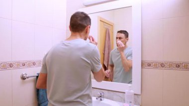 Adam, fayanslı bir banyoda pamuk pedleri ve yüz ürünleri kullanarak cilt temizleme rutinini gerçekleştiriyor.