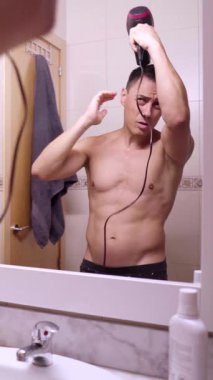 Gömleksiz bir adam banyo aynasının önünde saç kurutma makinesiyle saçını şekillendiriyor.