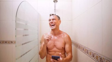 Mutlu adam duşta akıllı telefon kullanırken gülüyor.
