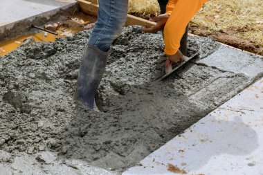 Mason worker is holding steel trowel and smoothing plastering new sidewalk on wet freshly poured concrete in construction site