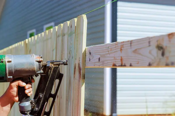 Man builds sections of nailing a wooden fence around his yard out planks using air nail gun