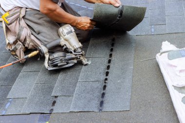 Roofer uses an air pneumatic nail gun to attach new asphalt bitumen shingles to roof clipart