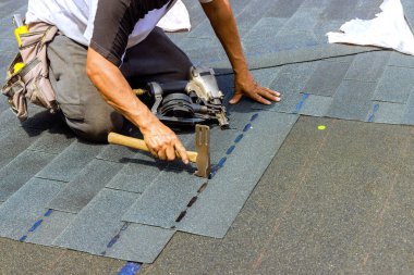 An installation worker using an air pneumatic nail gun installed new bitumen shingles on roof clipart