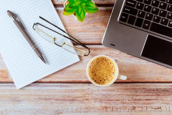 Çalışma alanı dizüstü bilgisayar, kahve, kalem ve defter ile bir ofis masasının üstünde.