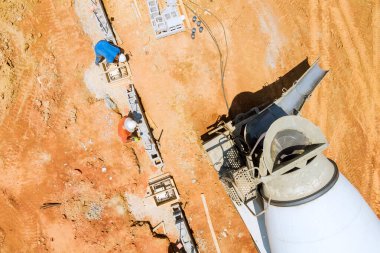 İşçi, büyük, kırmızımsı toprak renkli alanda inşaat temelleri üzerine beton dökmek için karıştırıcı kamyon kullanıyor.