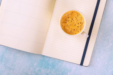 Cup of steaming coffee espresso sits next to an open notebook planner on light blue surface clipart