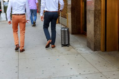 Two men in dress shirts, slacks stroll along busy city sidewalk, one pulling carry on suitcase, indicating travel professional meeting nearby. clipart