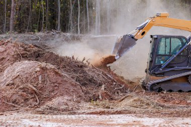 İnşaat buldozer makineleri aktif olarak ormandaki toprak kalıntılarını temizliyor. Kazıcı, gelecekteki gelişime hazırlanmak için malzeme toplarken toz bulutu yükseliyor..