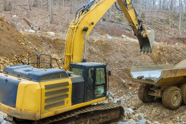 Large yellow excavator is loading dirt into nearby dump truck on construction site surrounded by machinery terrain. clipart