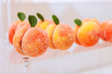 Delicate pastries resembling peaches are neatly arranged on transparent display dessert features fuzzy exterior with green leaf accents, creating an inviting appearance. clipart