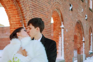 Newlywed couple embraces kisses under beautiful brick arches in snowy landscape their elegant attire contrasts with rustic surroundings, creating magical atmosphere. clipart