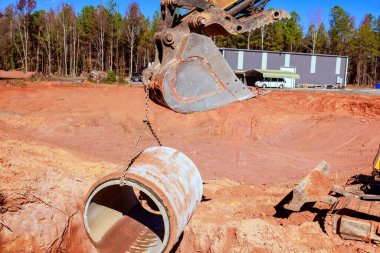 Excavator carefully lowers concrete pipe into freshly dug trench at construction site during installing drainage system clipart