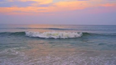 Altın, mor renkler gökyüzünü aydınlatıyor. Gün batımında sakin kumsallara doğru hafif dalgalar sıçrıyor..