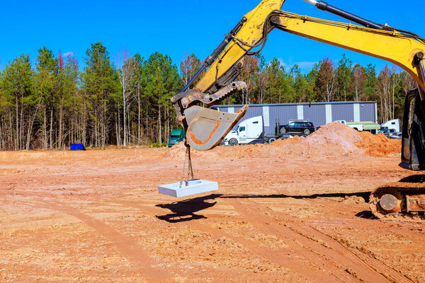Excavator lifts concrete slab at construction site with trucks storage buildings build works area