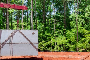 Crane lifts concrete septic tank onto construction site surrounded by lush green trees during construction new home clipart