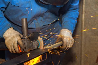 İşçi, metal parçayı bir açı öğütücüsüyle öğütüyor ve loş ışıklandırma atölyesinde kıvılcımlar oluşturuyor..