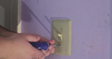 Bir insan mor bir duvarda tornavidayla ışık düğmesini tamir ediyor ve görevine odaklanıyor..