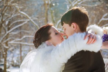 Gelin ve damat, karlı açık havada, ağaçlarla çevrili gelinin kürklü atkısını giyerek nazikçe kucaklaşır.,
