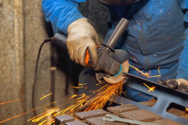 Metal işçisi çeliğin kenarlarını pürüzsüzleştirmek için öğütücü kullanırken, kıvılcımlar sönük atölyeyi aydınlatıyor..