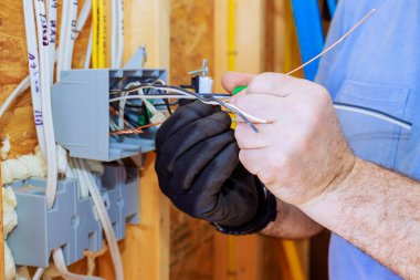 Bir elektrikçi kısmen inşa edilmiş evde eldiven giyerken bağlantı kutusundaki kabloları birbirine bağlıyor..
