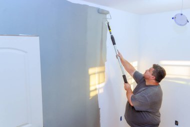 Müteahhit iyi aydınlatılmış bir odada merdivenin üstünde dikilirken duvarları tekerlekli sandalyeyle griye boyuyor..