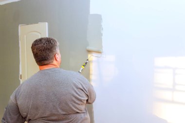 Müteahhit, iyi aydınlatılmış odada patenlerle duvara gri boya uyguluyor, ev geliştirme projesini gözler önüne seriyor.