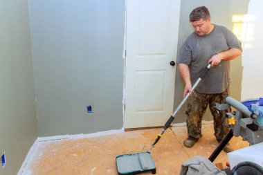 Adam araçlarla çevrili, odadaki açık gri duvarı boyamak için tekerlek kullanıyor..