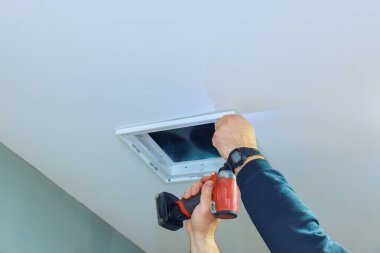 Tamirci, havalandırma kanalı kapağını iyi aydınlatılmış bir odanın tavanına yerleştirmeye çalışıyor..