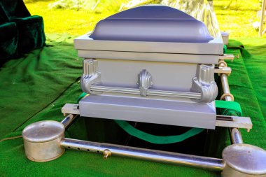 Gümüş tabut, yeni kazılmış mezarın üzerine yerleştirilir. Yeşil çimlerle, defin töreninde sessiz çevrelerle birlikte..