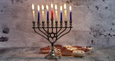 Dokuz yanan mumlu gümüş menorah, ön planda süslü hamur işleriyle dokulu bir yüzeye yerleştirilmiş kırmızı mum. Menorah dizaynında görünen Davut 'un Yıldızı, desenli beton duvar