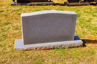 Gri granit mezar taşı huzur dolu bir mezarlıkta tek başına duruyor, yemyeşil çimlerin üzerindeki parlak gün ışığıyla aydınlanıyor..