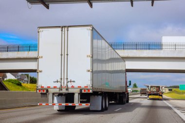 Büyük kamyonlar bulutlu gökyüzünün altında banliyöde üst geçitle yoğun Amerikan otoyolu boyunca seyahat ediyorlar..