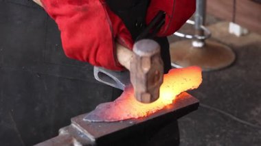 Bir zanaatkar, çekiç ve baltayla örs üzerinde kırmızı metali şekillendiriyor. Bu sırada usta demircilik yapıyor..