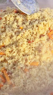 Etrafı buharla çevrili büyük bir tavada karıştırılmış havuçlu büyük miktarda pirinç çimenli arka planda pişirilmiş. Buhar yükselirken mutfak aletleri malzemeleri taşır