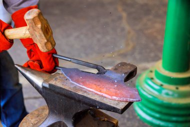 Bir zanaatkar, çekiç ve baltayla örs üzerinde kırmızı metali şekillendiriyor. Bu sırada usta demircilik yapıyor..