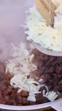 Et küpleri ve doğranmış soğan yakın çekim çimenlerin üzerinde büyük bir tencerede pişiriliyor, buhar yükseliyor ve delinmiş bir spatula malzeme ekliyor. Yemek pişirme süreci dinamik panelleme ile yakalandı