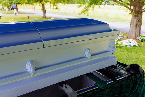 Белый гроб установлен на кладбище во время панихиды среди зеленых деревьев.