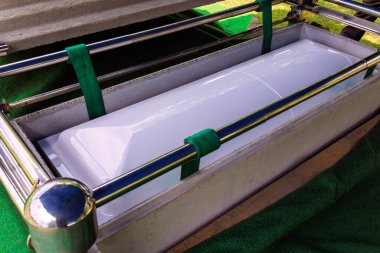 Metal çerçeveli beyaz tabut, sakin mezarlıktaki bir anıtta yeşil kumaşla çevrili.