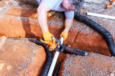 İnşaat işçisi yeni kazılan sipere drenaj kurarken PVC borusunu kesiyor.